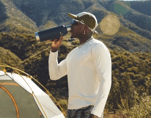 A man drinks from his water bottle in front of a tent.