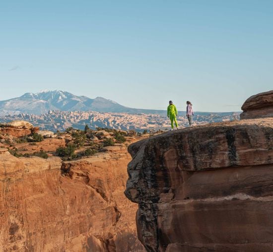 Two people stand on a red rock cliff, looking at the view.