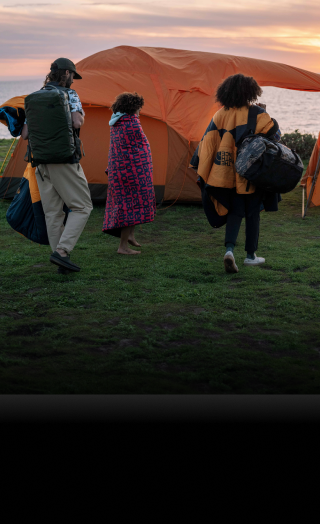 Three campers carry gear to their tent overlooking the ocean at sunset.