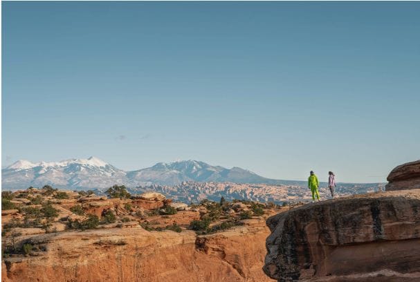 Two people stand on a red rock cliff, looking at the view.