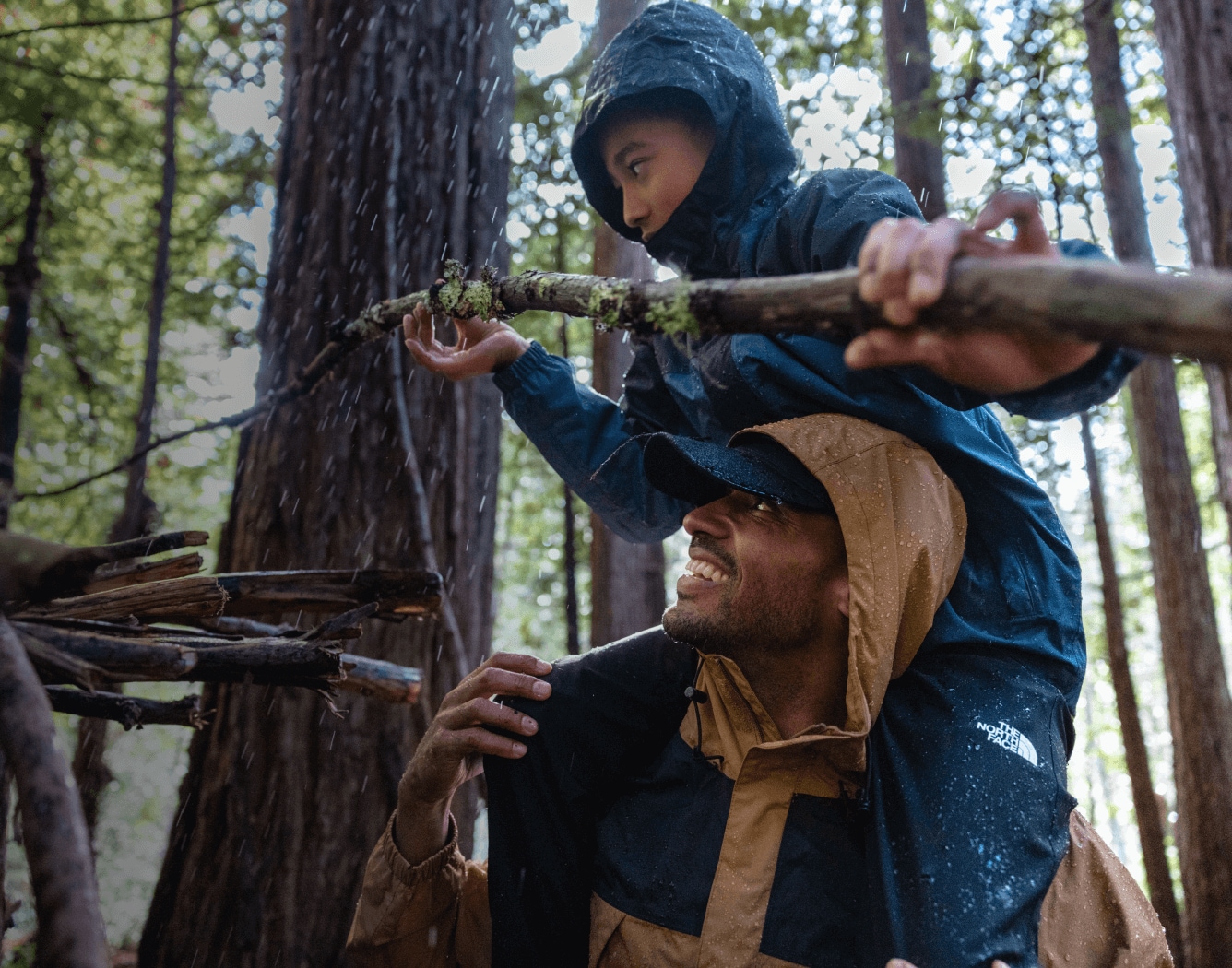 A man is giving a child a piggyback ride in a rainy forest while they wear rainwear from The North Face.