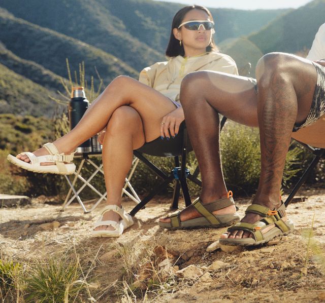 A man and woman wearing sandals from The North Face sit in camp chairs in a desert mountain setting.